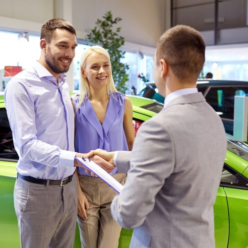auto business, car sale and people concept - happy couple with dealer shaking hands in auto show or salon